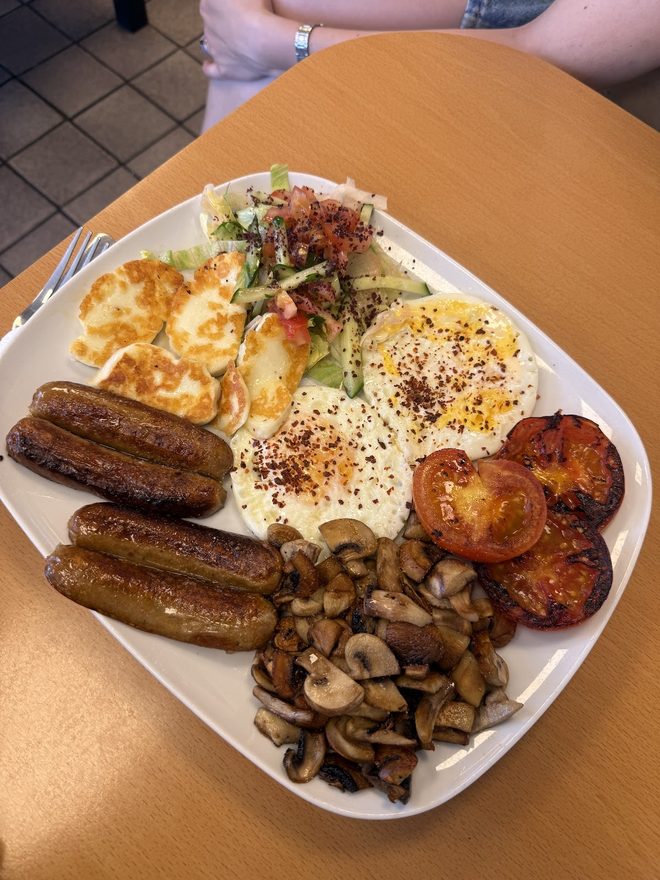 Sausages, eggs, halloumi, mushrooms and grilled tomatoes on a plate held over an outdoor table.